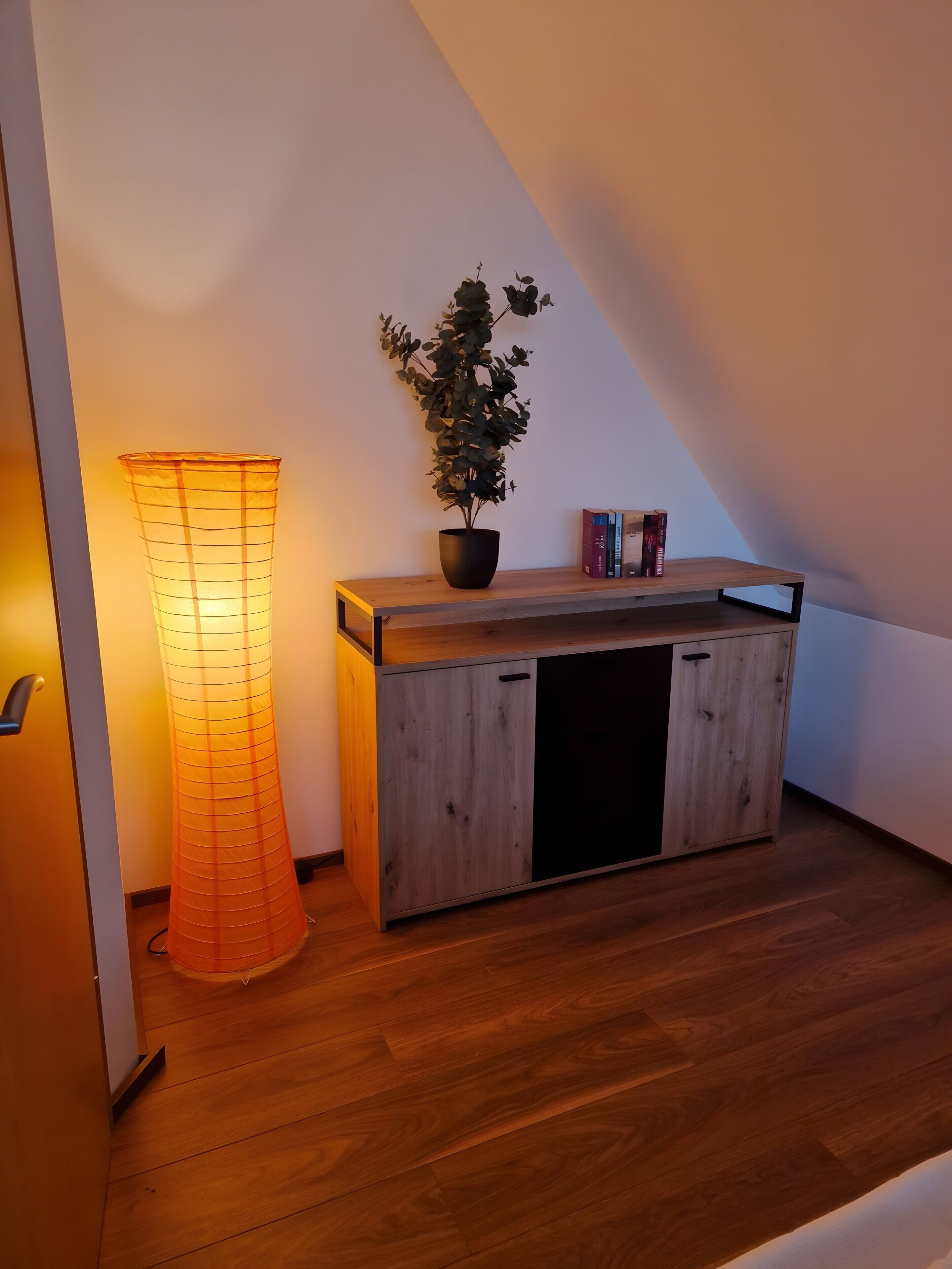 A wooden bench with a potted plant and books, a floor lamp, and a sloped ceiling.