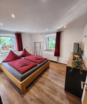 Un dormitorio moderno con una cama de madera, almohadas rojas, un pequeño escritorio con una planta y dos ventanas con cortinas.