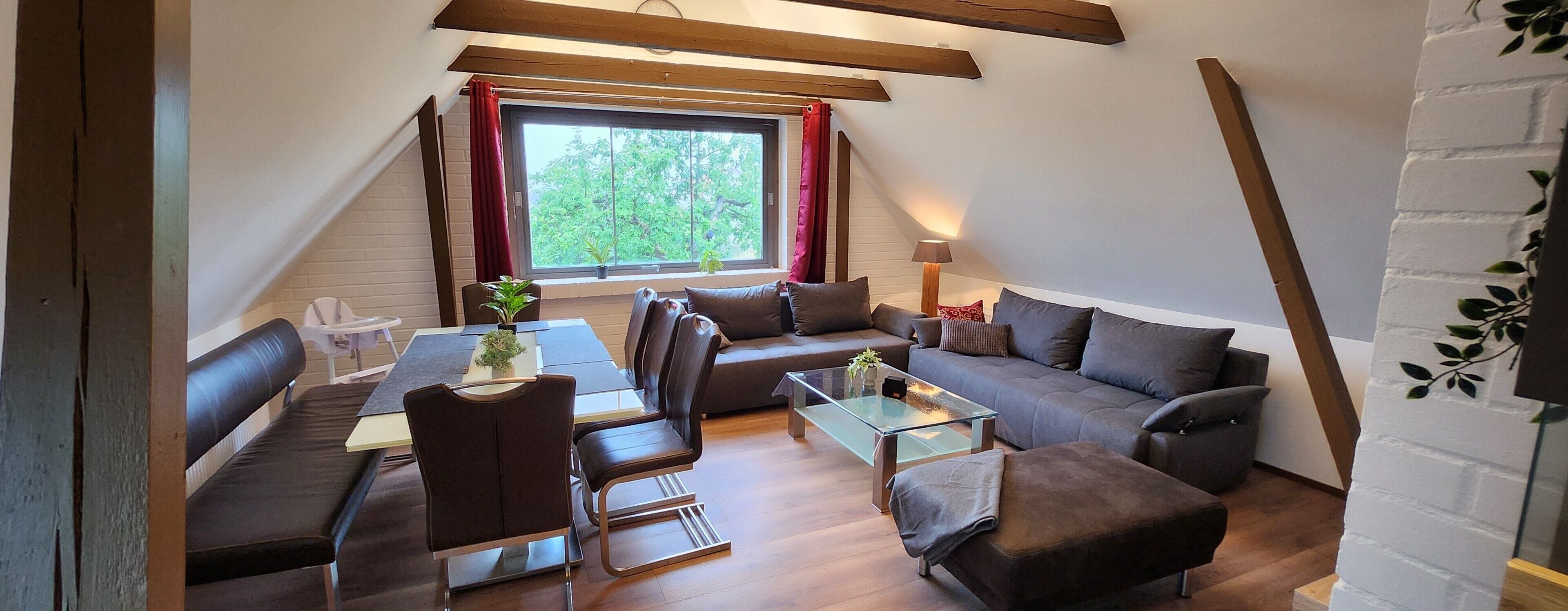 A cozy loft-style room with a dining table, sofa, and ottoman, a large window with a view of greenery, and exposed wooden beams.