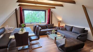 A cozy loft-style room with a dining table, sofa, and ottoman, a large window with a view of greenery, and exposed wooden beams.