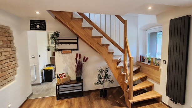 Un escalier en bois menant à un deuxième étage, un mur de briques à gauche, un radiateur à droite et une porte donnant sur l’extérieur.