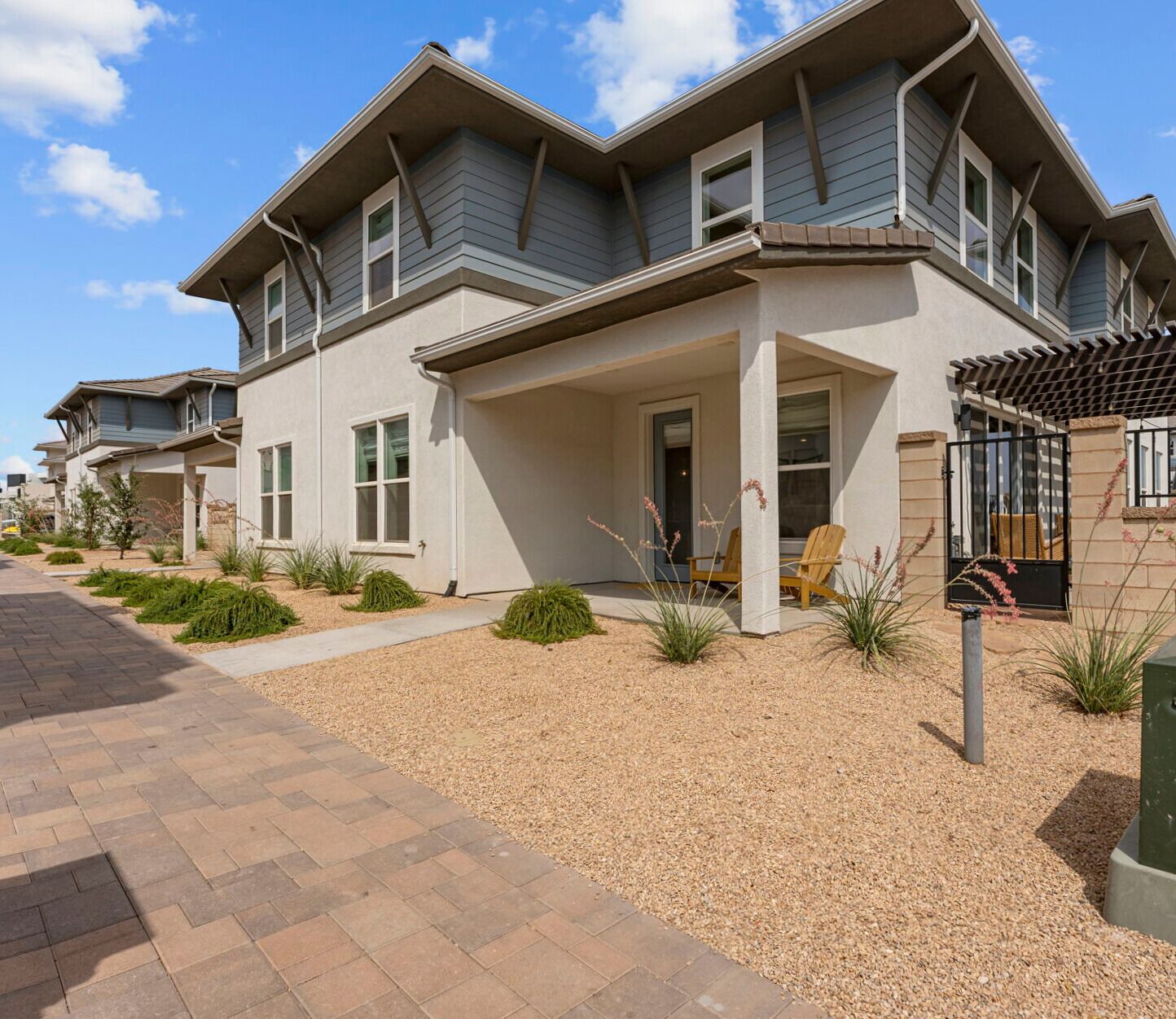 A modern two-story house with a covered porch, a small garden with various plants, and a paved driveway.