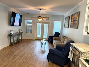 A living room with a ceiling fan, a television mounted on the wall, a sofa, a coffee table, and a kitchen area with a countertop. - Sandy Dreams, 2-bedroom dog-friendly duplex in Garden City w/fenced yard (Murrells Inlet)