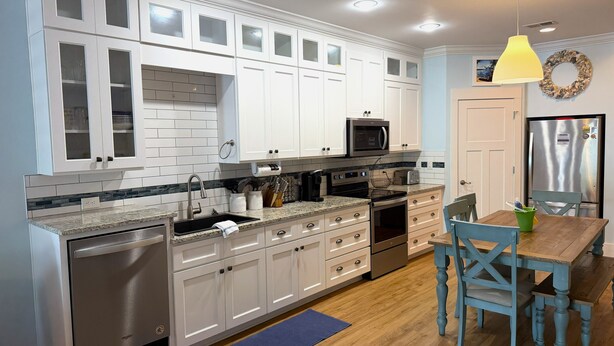 A modern kitchen with white cabinetry, stainless steel appliances, and a wooden dining table. - Sandy Dreams, 2-bedroom dog-friendly duplex in Garden City w/fenced yard (Murrells Inlet)