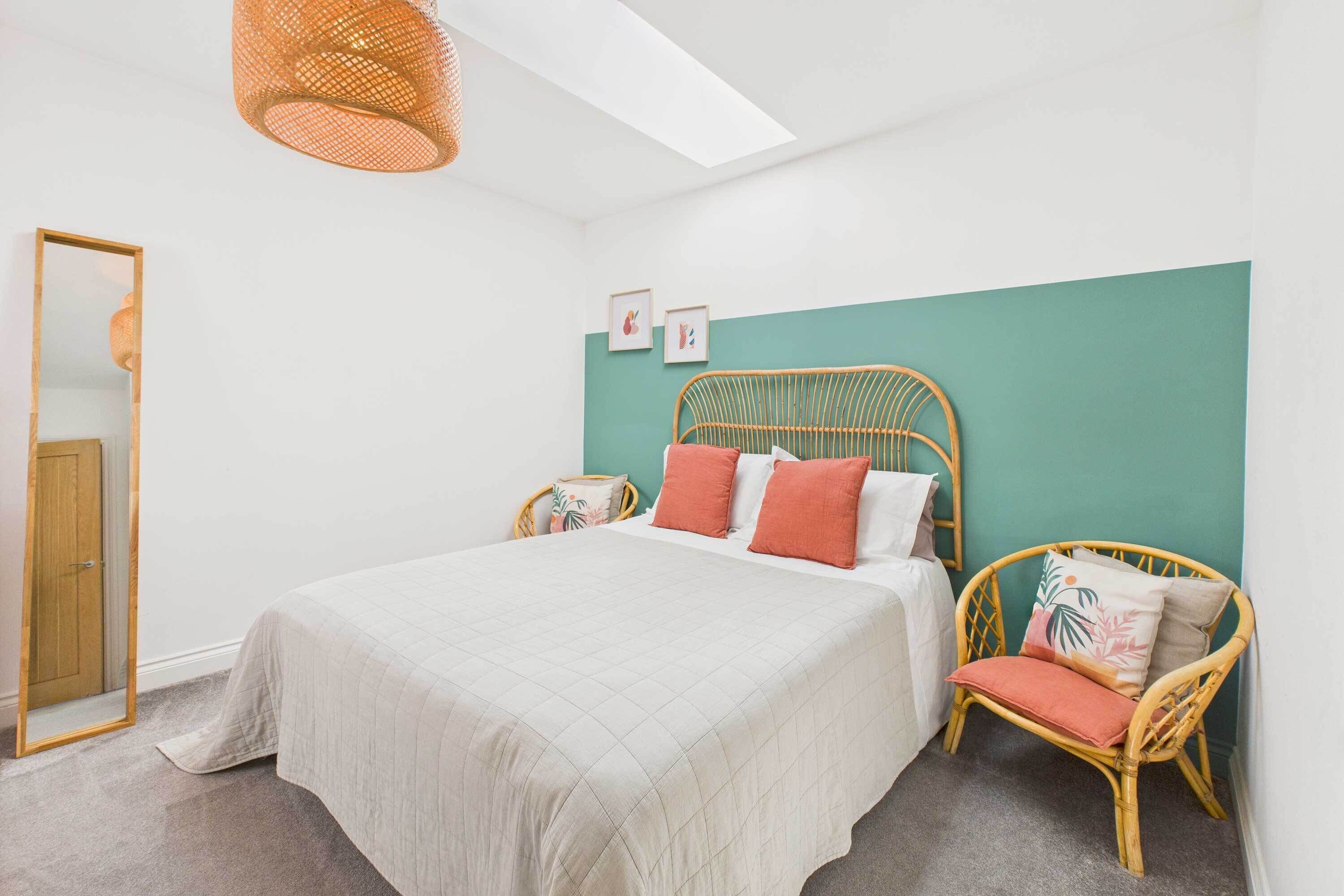 A bedroom with a bed, a wicker chair, a mirror, and a door.