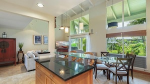 A spacious kitchen with a central island, wooden cabinets, and a dining area with chairs and a glass table. - Princeville Island Charm Townhome - Puamana 19C (Princeville)