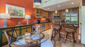 A modern kitchen with wooden cabinets, a glass dining table, and a large window. - Princeville Island Charm Townhome - Puamana 19C (Princeville)
