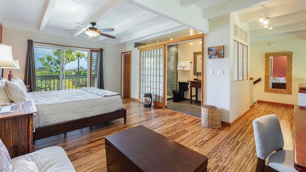 A spacious bedroom with a large bed, wooden flooring, a ceiling fan, and a view of greenery through a sliding door. - Princeville Island Charm Townhome - Puamana 19C (Princeville)