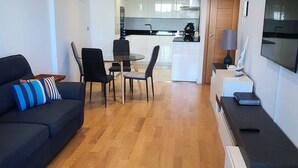 A modern living room with a dark blue sofa, a dining area with a round table and chairs, a kitchen with white cabinets, and a TV mounted on the wall. - One Bedroom Ocean Village Apt (Gibraltar)
