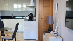 A modern kitchen with white cabinets, a dining area with a glass table and chairs, and a wooden door leading to another room. - One Bedroom Ocean Village Apt (Gibraltar)