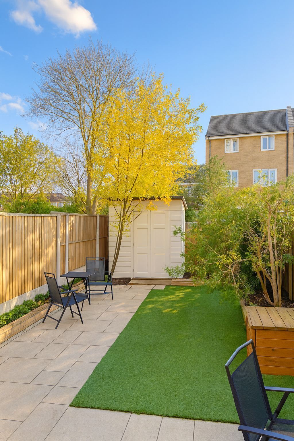 A backyard with a patio, artificial grass, a wooden fence, and a tree with yellow leaves.