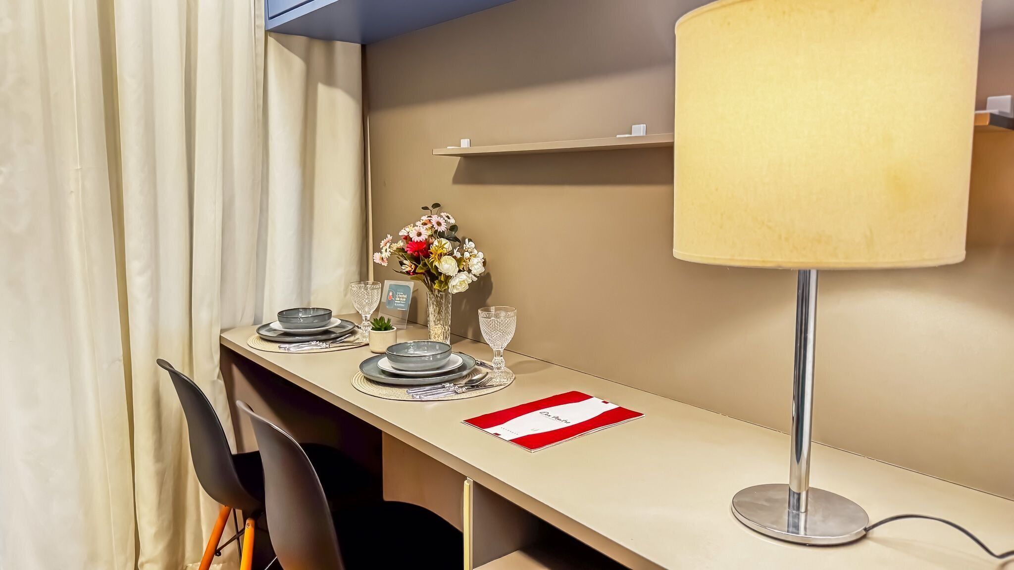 Ein ordentlich eingerichtetes Hotelzimmer mit Esstisch für zwei Personen, einer Blumenvase, einer Lampe und einem Regal mit Taschentuchbox.