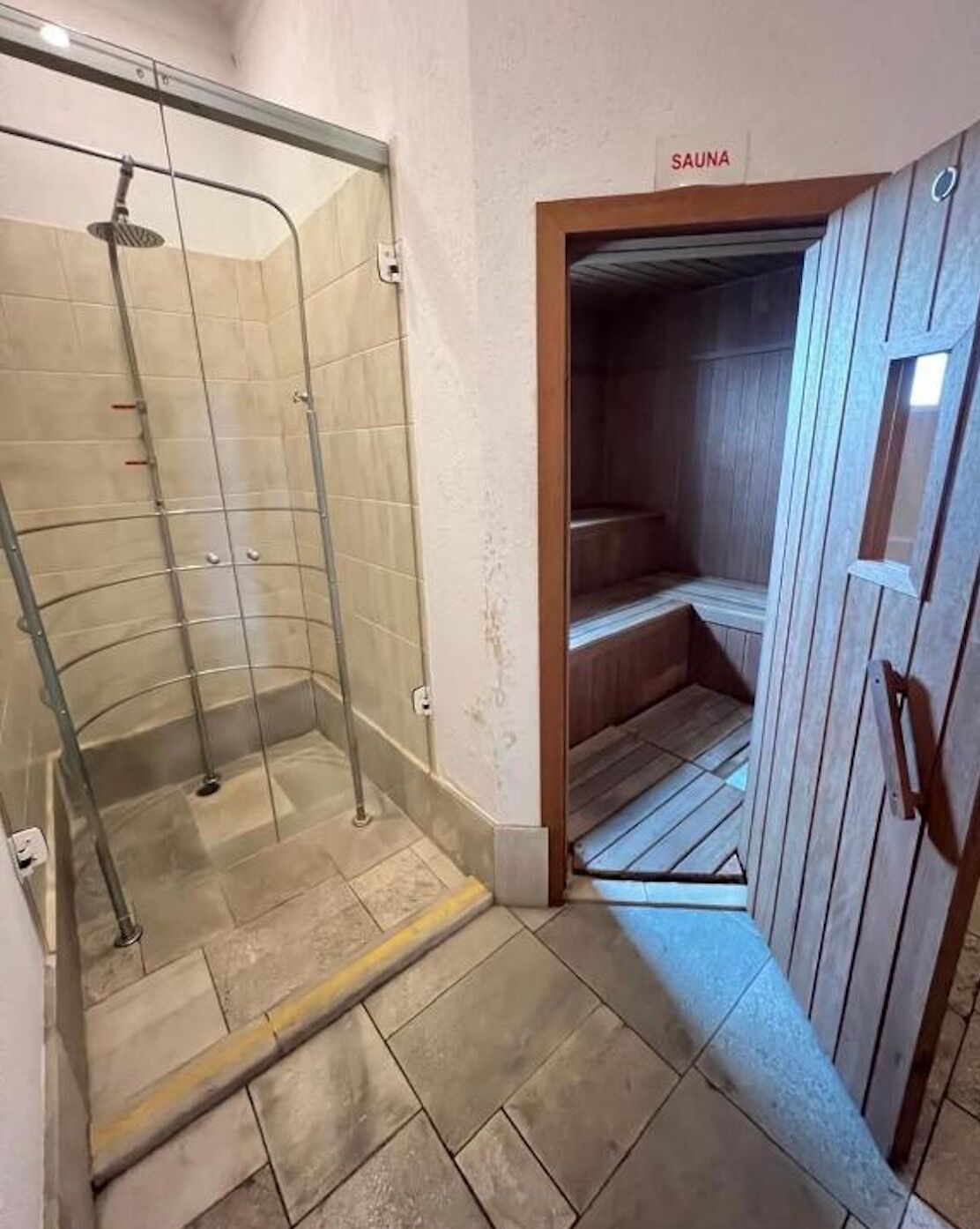Zona d'aixeta tancada amb vidre, sòl de rajola i entrada a la sauna amb parets de fusta.
