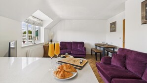 Un salon moderne avec un canapé violet, une table à manger avec des verres à vin et un coin petit-déjeuner avec des croissants et du jus d’orange.