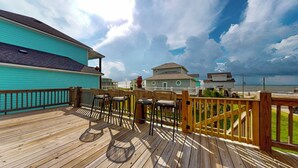 A wooden deck with outdoor seating, a turquoise building, and a view of houses and the ocean. - Casa de Tortuga: 4  BR, 3  BA House in Crystal Beach, Sleeps 12 (Bolivar Peninsula)