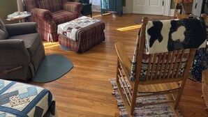 Un salon chaleureux avec un canapé à carreaux, une chaise en bois et un fauteuil bleu. Le sol est en bois, il y a un ventilateur de plafond et une fenêtre avec des rideaux.
