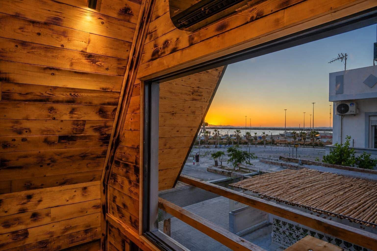 Ein Holzinnenraum mit einem großen Fenster mit Blick auf eine Küstenlandschaft bei Sonnenuntergang.