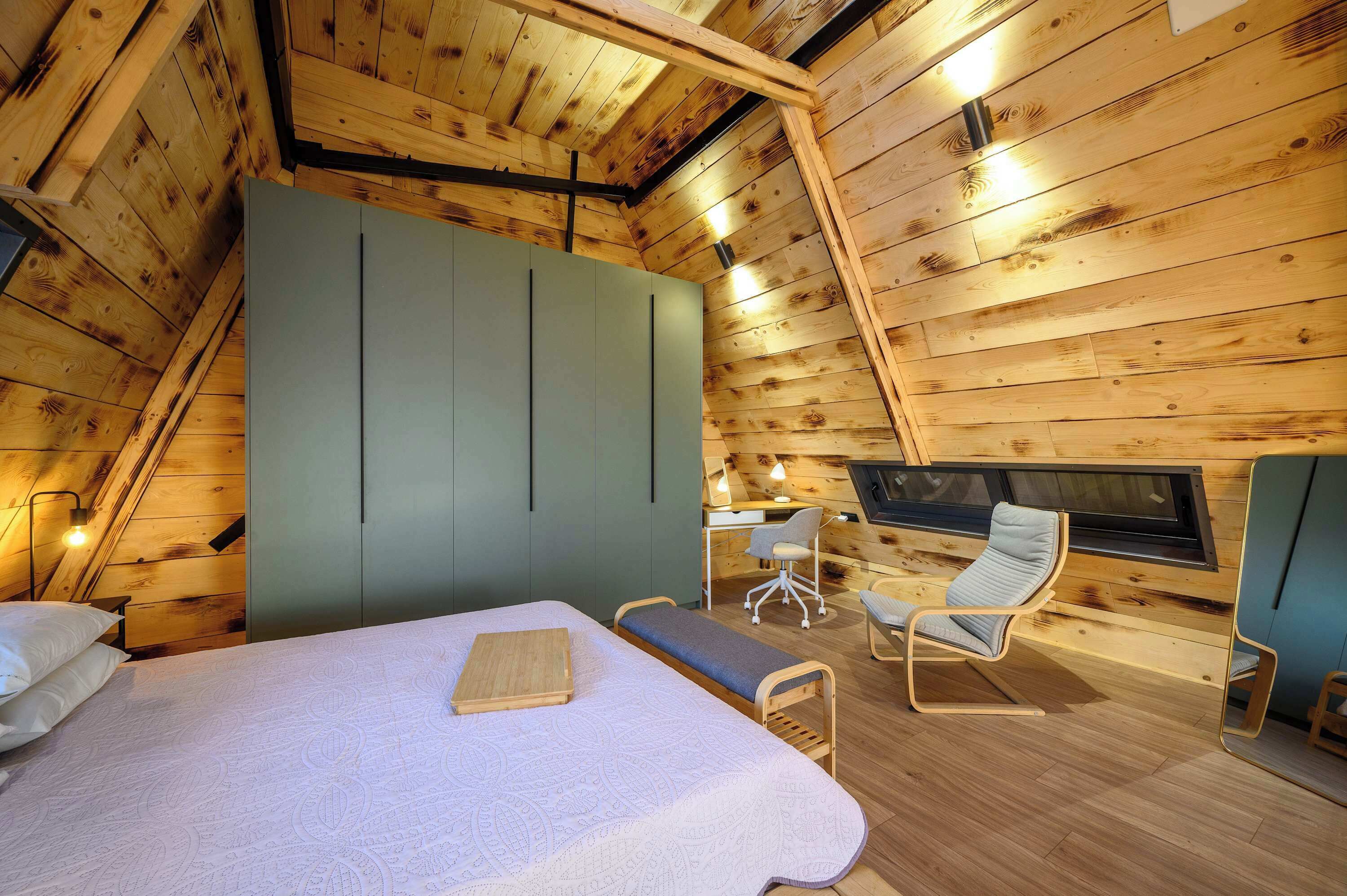 A cozy attic bedroom with a bed, a desk, and a chair, featuring wooden walls and a sloped ceiling.