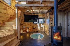 A wooden cabin interior with a central fireplace, a small circular window, and a bench along the left wall.