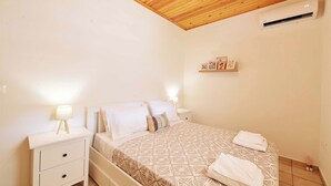 A bedroom with a wooden ceiling, a bed with a patterned bedspread, a bedside table with a lamp, and a wall-mounted air conditioner.