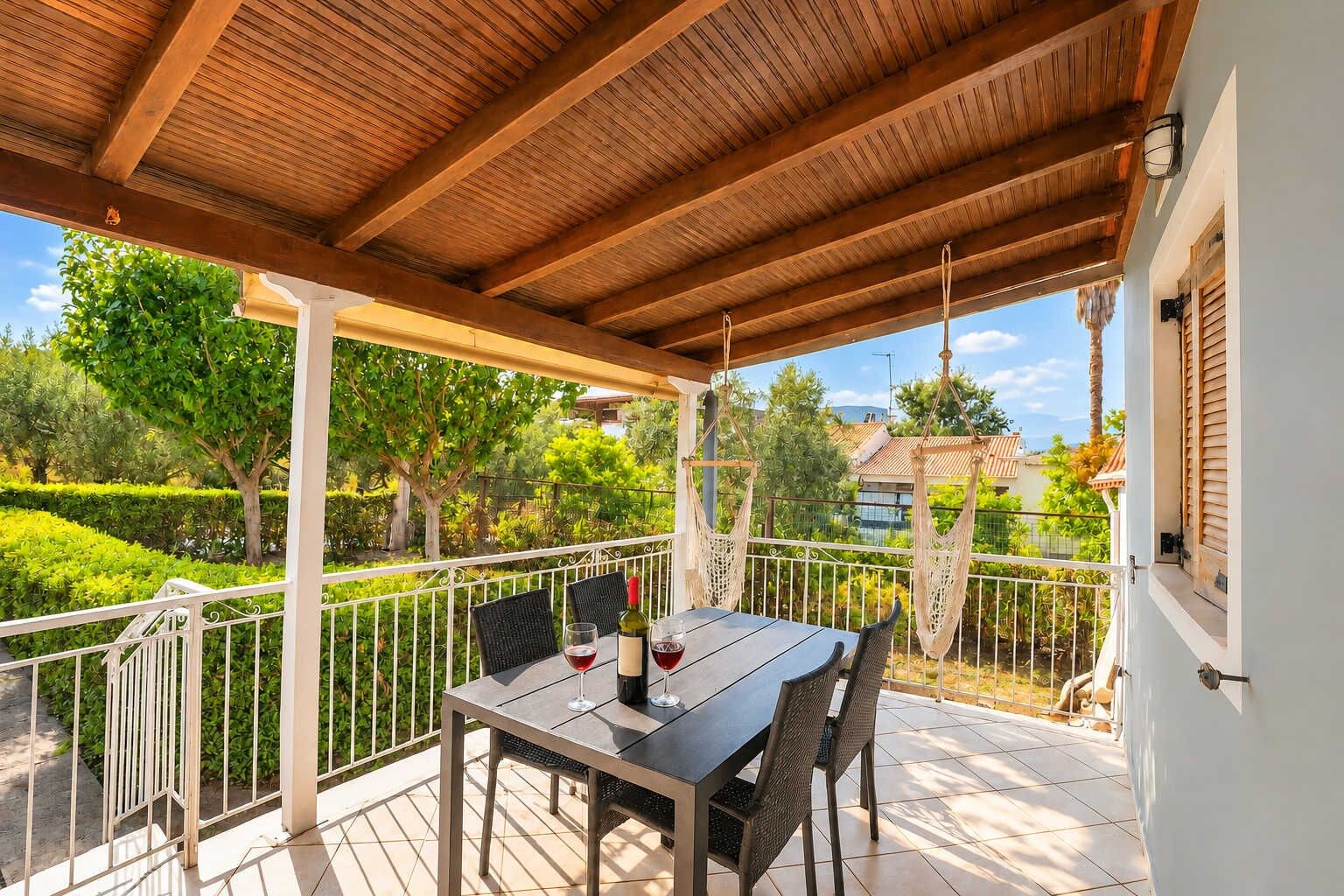 A covered outdoor patio with a table and chairs, a railing, and a view of the surrounding greenery and buildings.