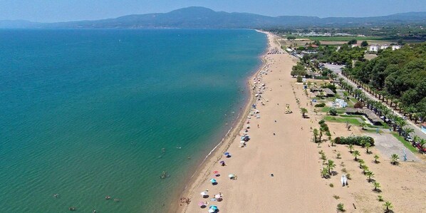 An aerial view of a beach with a long stretch of sand, a clear blue sea, and a line of trees and buildings along the shore.