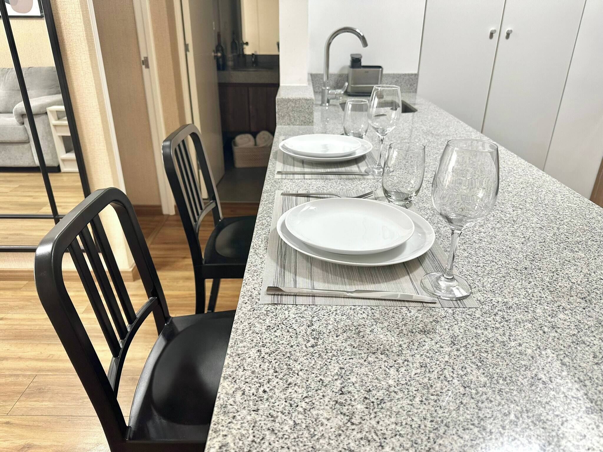 Une cuisine dotée d’un plan de travail en granit, de deux chaises noires, d’une table à manger mise avec des assiettes et des verres à vin, et d’un évier avec un robinet.