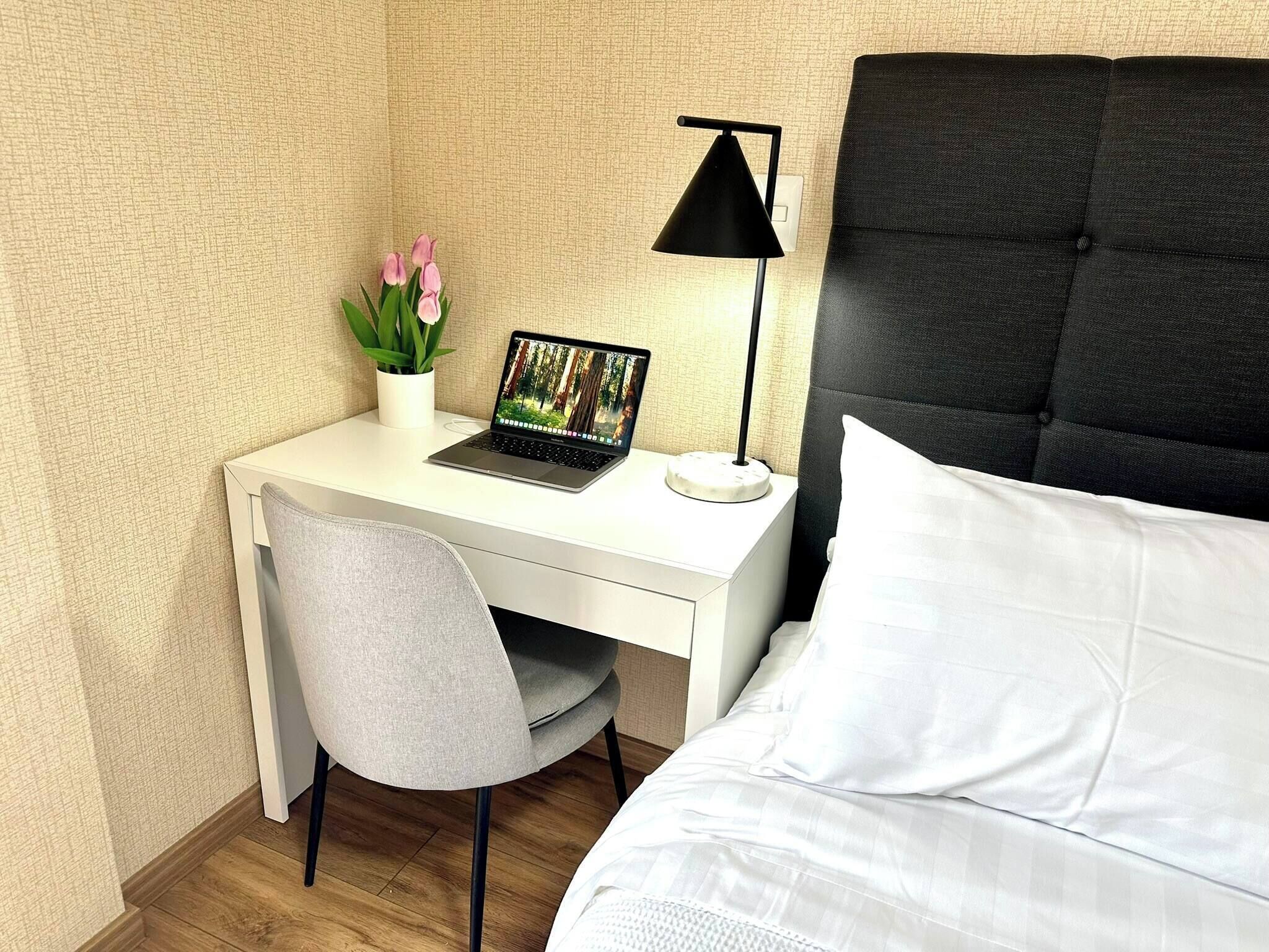 Une chambre d’hôtel avec un bureau blanc, un ordinateur portable, une lampe noire, une chaise grise et un lit avec une literie blanche.