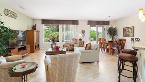 A spacious living room with a sofa, armchairs, a coffee table, and a TV. There is a dining area with a table and chairs.