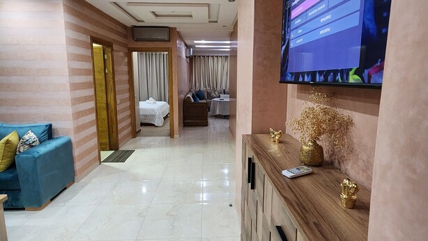 Ein Hotelzimmer mit einem großen Flachbildfernseher an der Wand, einem Holzkonsolentisch mit Dekoration, einem blauen Sofa mit Kissen und einem Bett mit weißer Bettdecke.