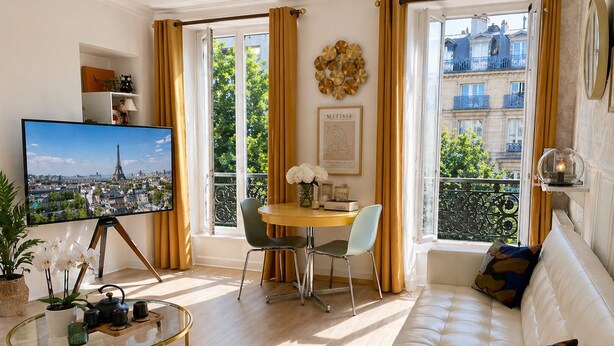 Ein modernes Wohnzimmer mit einer Couch, einem Esstisch mit Stühlen, einem Fernseher, der eine Stadtansicht zeigt, und großen Fenstern mit Vorhängen.
