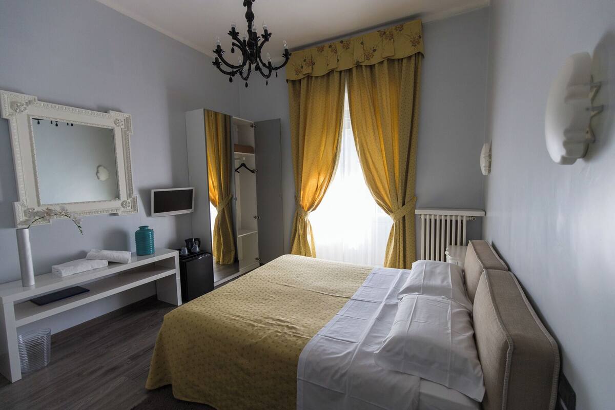 A bedroom with a bed, a bedside table, a mirror, a TV, and a window with curtains.