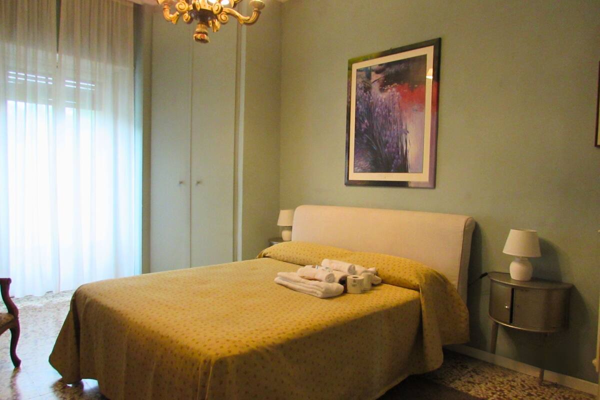 A hotel room with a bed, a nightstand, a lamp, a painting on the wall, and a chandelier.
