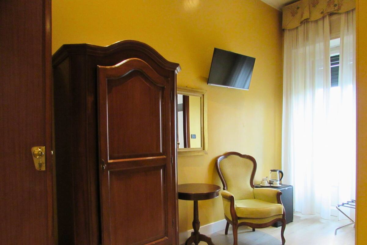 A room with a wooden door, a round table with a chair, a flat-screen TV mounted on the wall, and a window with curtains.
