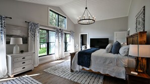 A bedroom with a large bed, a TV, a wooden dresser, and a window with curtains.