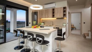 A modern kitchen with a central island, bar stools, and a view through a glass door. - Terra Magna - Alba Villa (Rhodes)