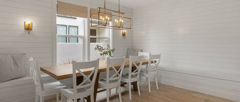 A dining area with a wooden table and white chairs, a hanging light fixture, and a window with blinds.