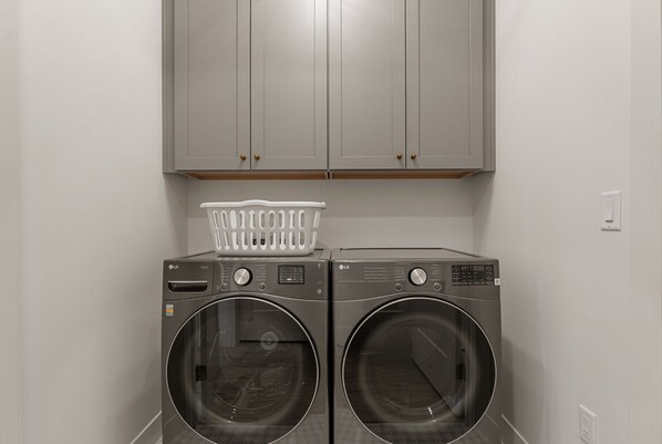 A laundry area with two front-loading washers and dryers, a white laundry basket, and grey cabinets above.