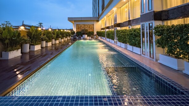 A swimming pool with a tiled edge, surrounded by potted plants and a building with large windows and a modern design. - Golden Empire Suvarnabhumi Hotel (Bang Phli)