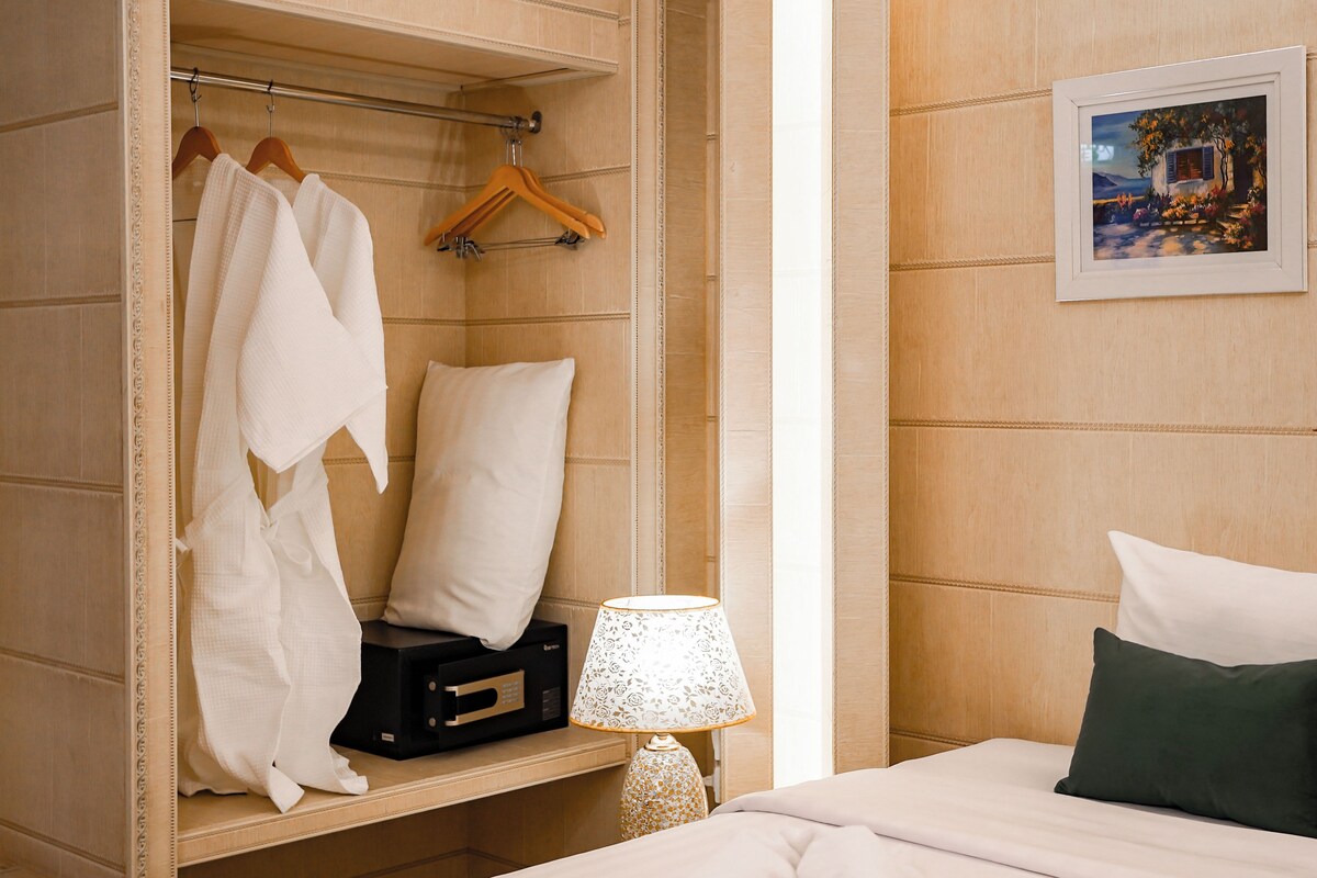 Habitación de hotel con vestidor, cama con ropa de cama blanca, almohada verde, lámpara y un cuadro en la pared.
