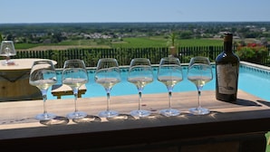 A wooden table with five wine glasses and a bottle of wine, overlooking a swimming pool and a scenic landscape.