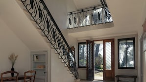 A spacious room with a high ceiling, a staircase with ornate black railings, and large windows allowing natural light.