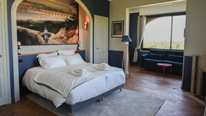 A bedroom with a large bed, a painting above the bed, a sofa, and a view through a window.