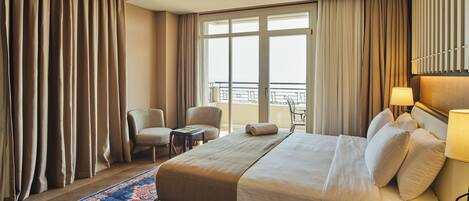A hotel room with a large bed, two armchairs, a small table, a balcony with a railing, and a view of the city.