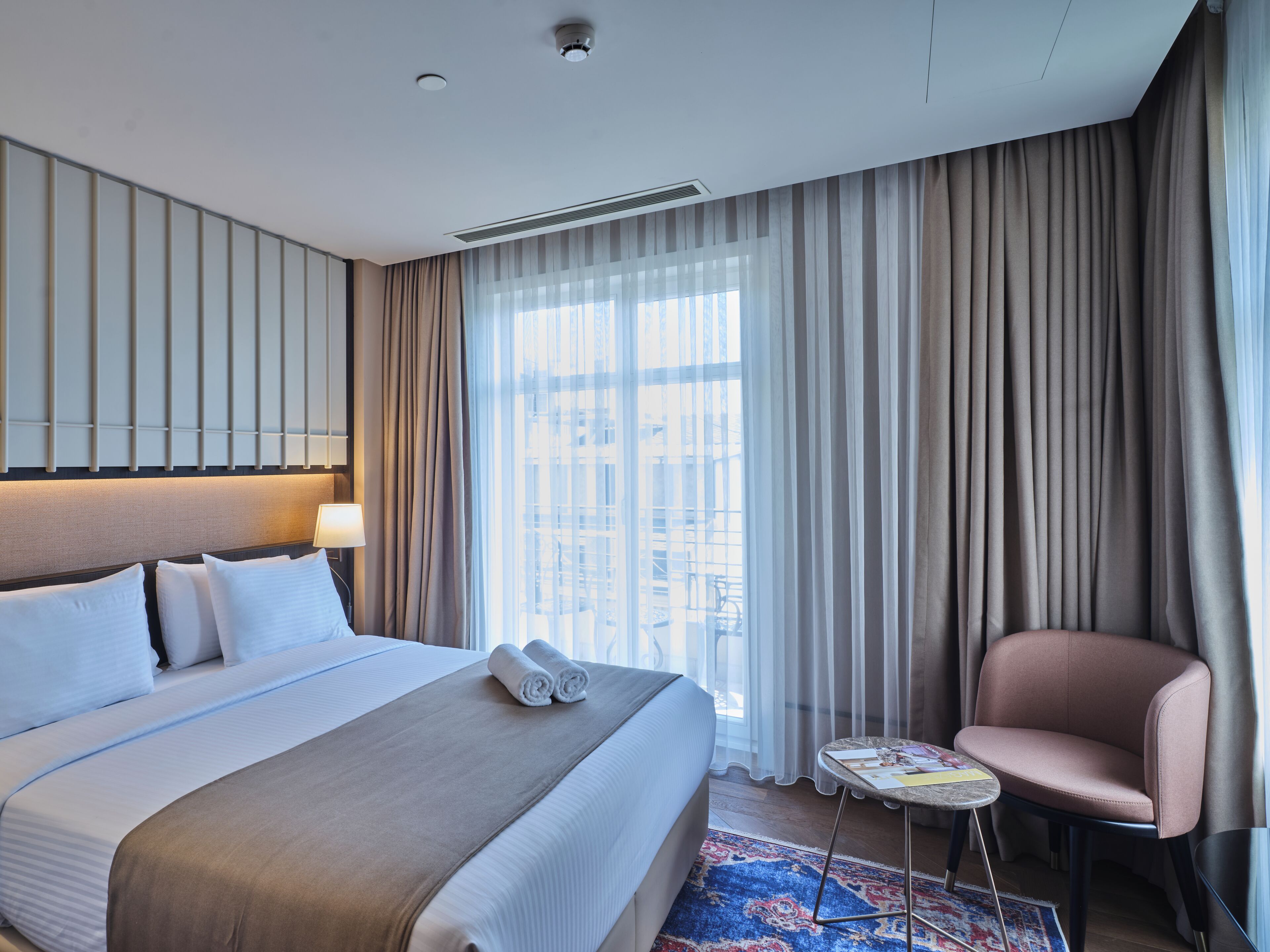 A hotel room with a large bed, a chair, a small table, and a view of the city through sheer curtains.