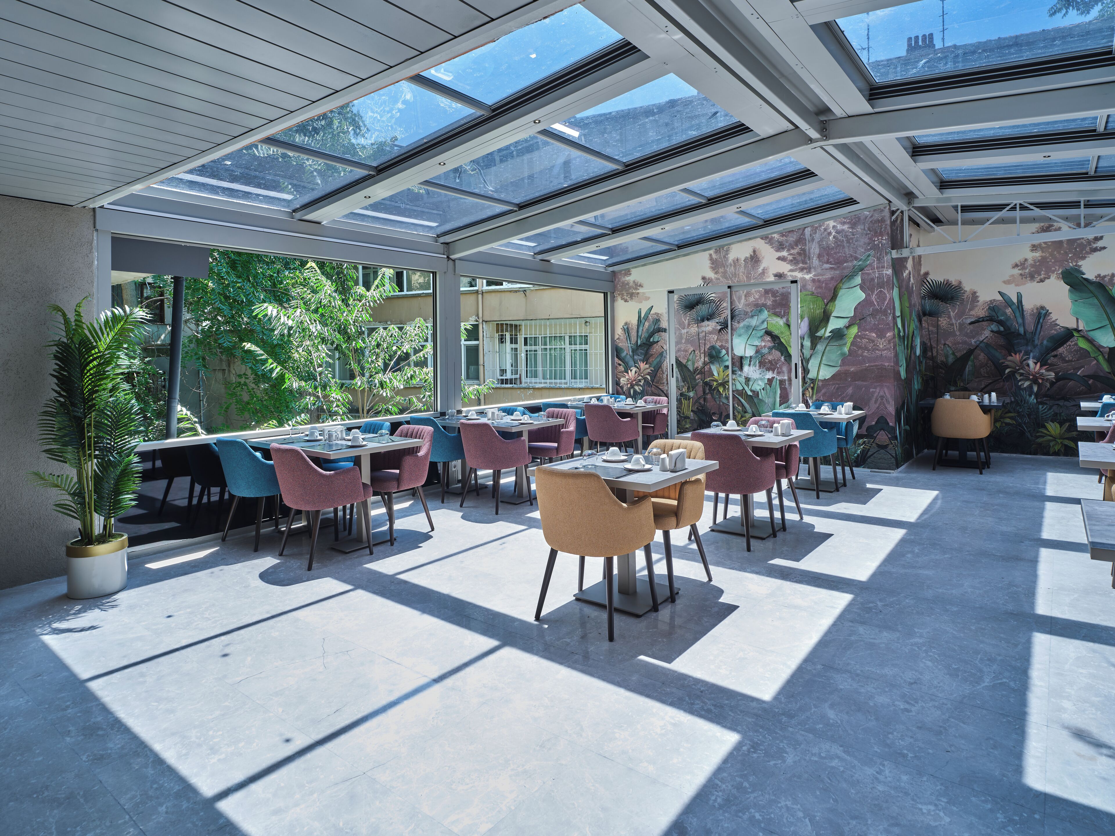 An outdoor dining area with tables and chairs, large windows, and a mural of tropical plants.