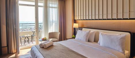A hotel room with a large bed, a small table with a fruit bowl, a chair, and a view of the sea through a balcony.