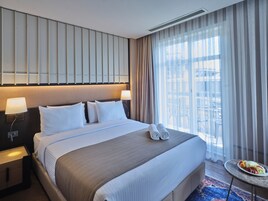 Una habitación de hotel con una cama grande, dos lámparas de noche, un cabecero con paneles verticales, una ventana con cortinas traslúcidas y una mesita con fruta.