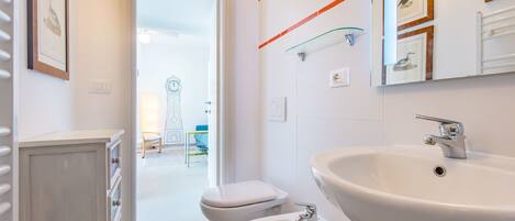 A modern bathroom with a large sink, bidet, and toilet. There is a glass shelf above the sink and a mirror above the toilet.