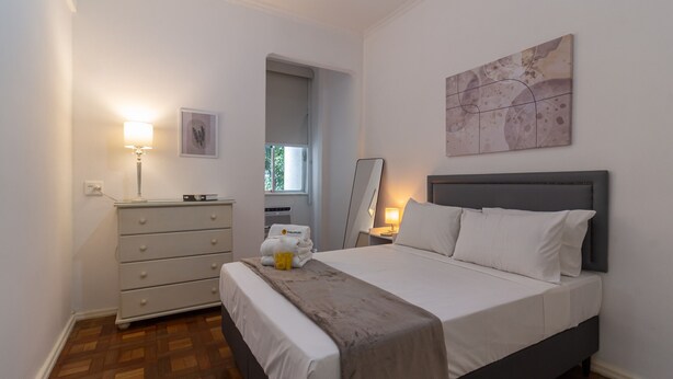 A bedroom with a bed, a dresser, a lamp, a window, and a painting on the wall. - Comfort in Copacabana, 9 minutes from the beach | CJ202 (Rio de Janeiro)